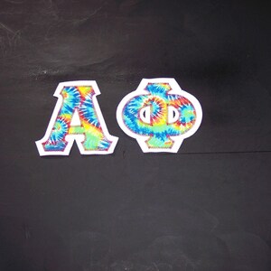 May include: Two embroidered patches featuring the Greek letters Alpha and Phi. The letters are filled with a vibrant tie-dye pattern in shades of blue, yellow, and green. Each letter is outlined in white with a decorative stitch.