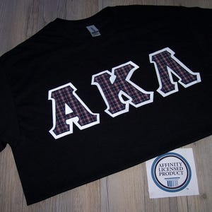 May include: Black t-shirt with the letters "AKA" in a collegiate style. The letters are white with a plaid pattern in shades of pink, purple, and black. A sticker with a blue and white design is in the lower right corner.