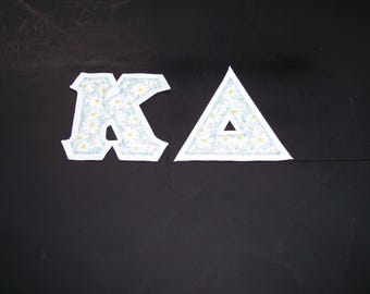 May include: Two fabric letters, K and Delta, featuring a light blue background with a white daisy floral pattern. Each letter has a white stitched border edge, creating a distinct outline against the solid black background of the image.