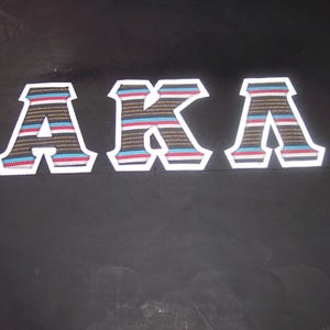 May include: The letters "AKA" are displayed in a row. Each letter is a patch with a white border and a striped design in brown, blue, and red. The background is a solid black.