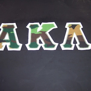 May include: Three fabric letters spelling "AKA" in a camouflage pattern. The letters are outlined in white with a green stitched border, set against a dark background. The letters are in a collegiate style.