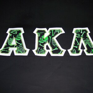May include: Black background with the letters "AKA" in white, outlined with a green zig-zag stitch. The letters are filled with a black pattern of green skeletons.