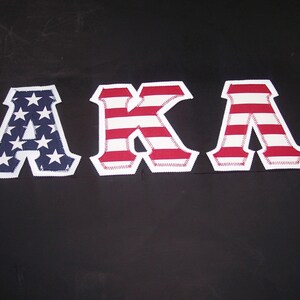 May include: Three fabric letters spelling "AKA" are displayed against a black background. The letter "A" on the left features a blue field with white stars, while the "K" and the second "A" have red and white stripes.