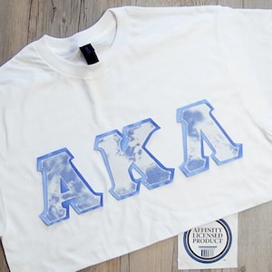 May include: White t-shirt with the letters "AKA" in a light blue, cloud-like design. The shirt has a crew neck and short sleeves. A small, round sticker with text and a barcode is visible.
