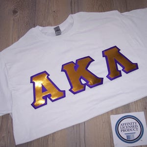 May include: White t-shirt with the letters "AKA" in gold and purple. The letters are large and bold, with a gold fill and purple outline. A small sticker with text is in the bottom right corner. The shirt is laying on a wooden surface.
