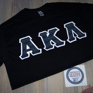 May include: Black t-shirt with the letters "AKA" in a white outline with a plaid pattern. The shirt is on a wooden surface. A sticker with the text "Affinity Licensed Product" is visible.