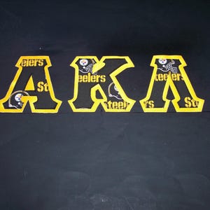 May include: Black and gold embroidered patches spelling out "AKA" with the Pittsburgh Steelers logo. The letters are outlined in gold with a black interior. The Steelers logo is visible within the letters.