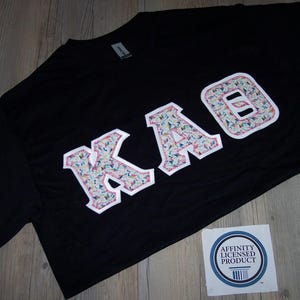 May include: Black t-shirt with the letters KAO in a floral pattern with a white border. The shirt has a tag on the upper back and a sticker with the text "Affinity Licensed Product" on the bottom right.