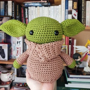 May include: A green crochet doll with large pointed ears and black plastic eyes, wearing a tan hooded robe. The doll has a round head and a stout body, with green hands visible at the ends of the sleeves. The texture is consistent throughout.