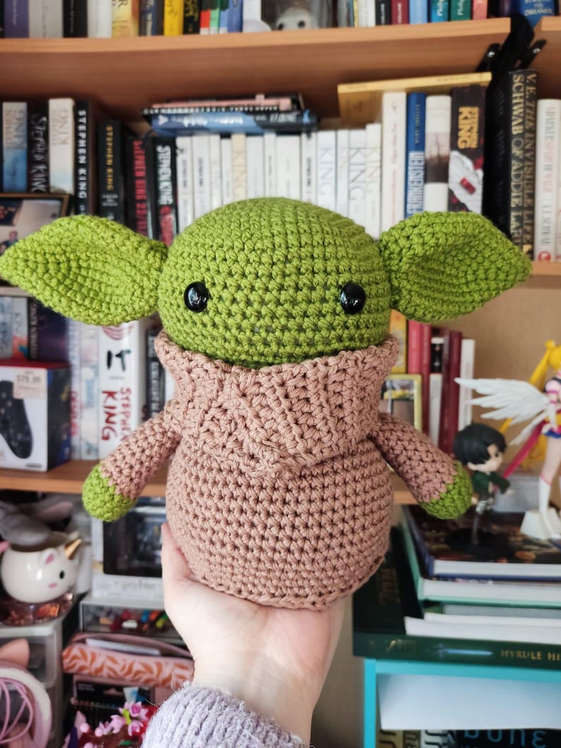 May include: A green crochet doll with large pointed ears and black plastic eyes, wearing a tan hooded robe. The doll has a round head and a stout body, with green hands visible at the ends of the sleeves. The texture is consistent throughout.