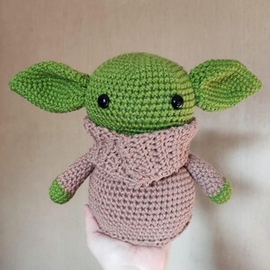 May include: A crochet plush toy featuring a round green head with large pointed ears, black plastic eyes, and a tan-colored hooded robe. The figure has a rounded body and small green-tipped arms, crafted with visible yarn stitches throughout.