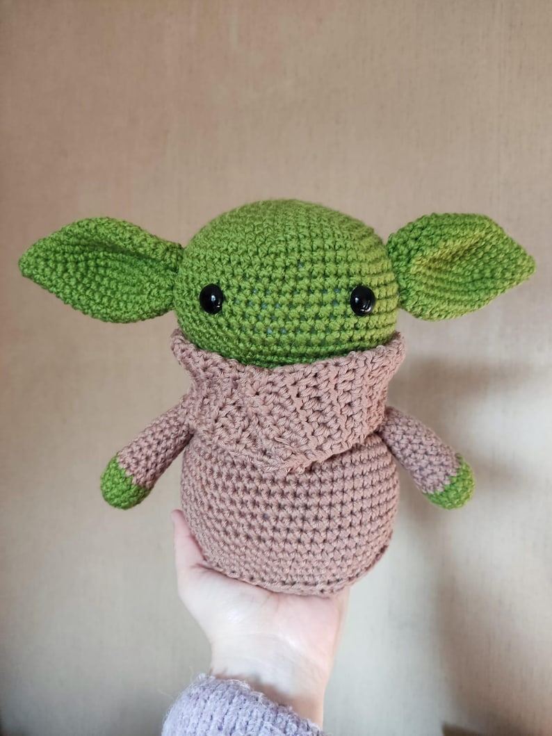 May include: A crochet plush toy featuring a round green head with large pointed ears, black plastic eyes, and a tan-colored hooded robe. The figure has a rounded body and small green-tipped arms, crafted with visible yarn stitches throughout.