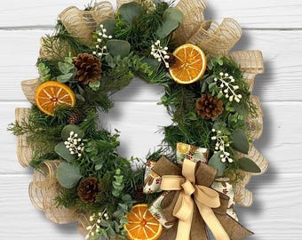 Puede incluir: Una corona decorativa con follaje verde, piñas y rodajas de naranja secas, adornada con una cinta de arpillera. Ideal para la decoración interior de temporada, con una estética rústica y natural.