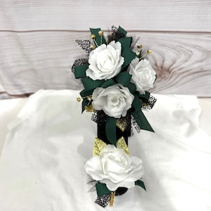 May include: A white flower corsage with green leaves and gold accents. The corsage is tied with black ribbon and is perfect for a special occasion.