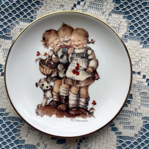 May include: A white decorative plate with a gold rim featuring a Hummel illustration of three children laughing and hugging, with a small dog and a basket. The plate has a glossy finish.