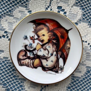 May include: A small, white decorative plate with a gold rim. The plate features a vintage illustration of a child under a red umbrella. The child is surrounded by flowers. The artist's signature, M.J. Hummel, is visible.