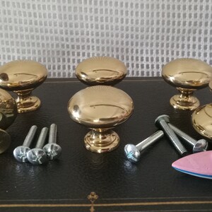 May include: A set of five polished gold-tone metal cabinet knobs with a smooth, rounded top and a tapered base. The set includes several silver-tone metal mounting screws and a small pink and blue toy boat figurine placed among the hardware.