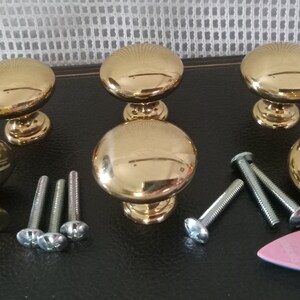 May include: A set of five polished gold-tone metal cabinet knobs with a smooth, rounded mushroom-shaped top and a tapered base. The set includes several silver-tone metal mounting screws and a small pink plastic toy boat figurine.