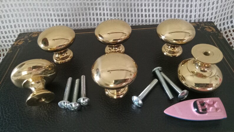 May include: A set of five polished gold-tone metal cabinet knobs with a smooth, rounded mushroom-shaped top and a tapered base. The set includes several silver-tone metal mounting screws and a small pink plastic toy boat figurine.