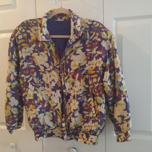 May include: A long-sleeve zip-up jacket featuring a vibrant floral pattern with white, yellow, and green flowers against a deep purple background. The jacket includes a high collar, a full-length front zipper, and elasticized cuffs and hemline.