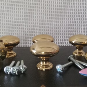 May include: A set of five polished gold-tone metal cabinet knobs with a smooth, rounded mushroom-shaped top and a circular base. The set includes several silver-tone metal mounting screws and a small pink and blue toy boat figurine.