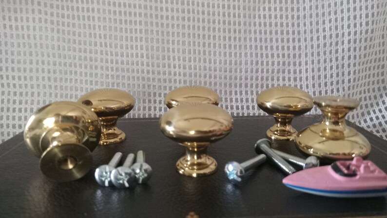 May include: A set of five polished gold-tone metal cabinet knobs with a smooth, rounded mushroom-shaped top and a circular base. The set includes several silver-tone metal mounting screws and a small pink and blue toy boat figurine.