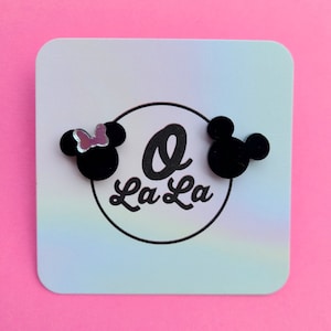 May include: A pair of black acrylic earrings featuring a Mickey Mouse and Minnie Mouse design. The Minnie Mouse earring has a pink bow. The earrings are displayed on a white card with the text "La La" in black.