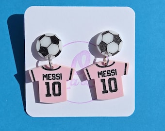 Puede incluir: Un par de pendientes de camiseta de fútbol rosa y blanca con un abalorio de balón de fútbol negro. La camiseta tiene el número 10 y la palabra "Messi" impresa en ella.