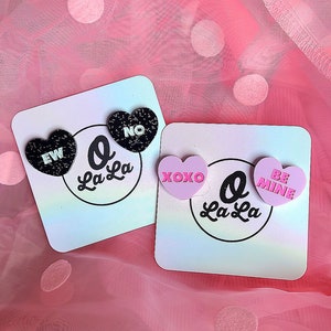 May include: Two pairs of heart-shaped earrings on cards. One set features black glitter hearts with "EW" and "NO" in white. The other has pink hearts with "XOXO" and "BE MINE" in pink. Each card is printed with "O La La".
