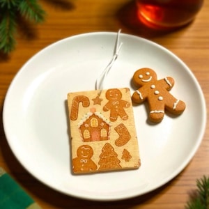 May include: A white plate holds a gingerbread man and a rectangular gingerbread ornament with Christmas-themed designs. The ornament has a house, candy cane, and other festive shapes. A glass of amber liquid sits nearby.