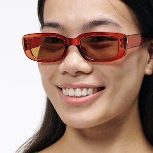 May include: A woman wearing brown rectangular sunglasses with a light brown frame. She is smiling and looking to the side.