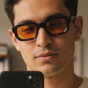 May include: Black rectangular eyeglasses with orange-tinted lenses. The glasses have a thick frame and are worn by a person holding a black smartphone. The person is wearing a cream-colored sweater. A bookshelf is visible in the background.
