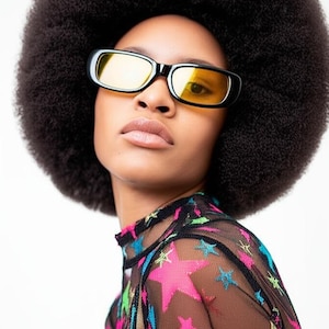May include: A person with an afro hairstyle wearing black sunglasses with yellow lenses and a black mesh top with colorful stars.