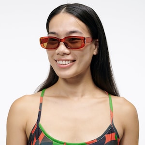 May include: A woman with long black hair wearing a red and green patterned tank top and brown rectangular sunglasses.