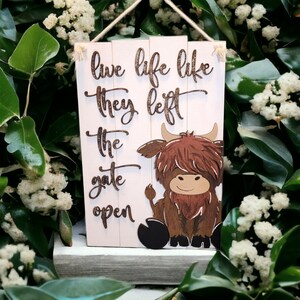 May include: A white wooden sign with the text "live life like they left the gate open" in brown lettering. A brown Highland cow with a white face is sitting on the sign.
