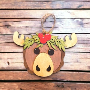 May include: Wooden Christmas ornament shaped like a moose head with a wreath of green leaves and red berries. The moose has a brown face with a light brown nose and large brown eyes.