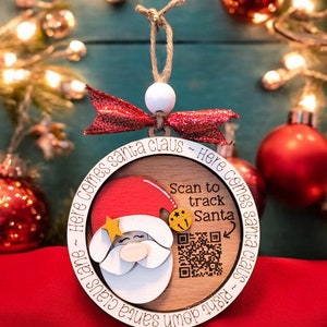May include: A round wooden Christmas ornament with a Santa Claus illustration and a QR code. The ornament reads "Here comes Santa Claus - Right down Santa Claus" and "Scan to track Santa".