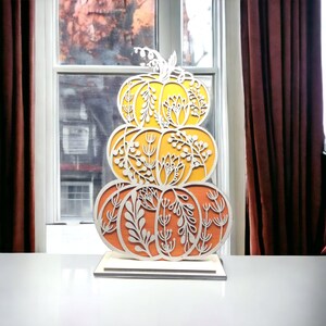May include: A wooden cutout of three stacked pumpkins with intricate floral designs. The pumpkins are painted in shades of orange and yellow.