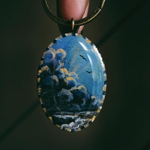 Peut inclure: Un porte-clés doré avec un charme rond de forme ovale représentant une scène peinte à la main d'un ciel bleu avec des nuages blancs et quelques oiseaux volant au-dessus d'une mer bleu foncé avec des vagues blanches.