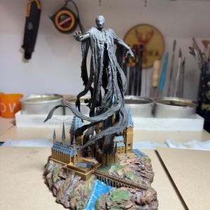 May include: A detailed figurine of a cloaked figure with outstretched arms, atop a miniature Hogwarts castle. The statue is predominantly gray, with the castle featuring tan, blue, and brown accents. A blue river flows through the base.