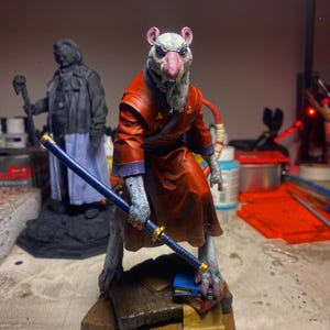 May include: A detailed figurine of a rat-like character in a red robe, holding a blue katana. The figure stands on a wooden base with books. Another statue is in the background. The figure has a pink nose and gray fur.
