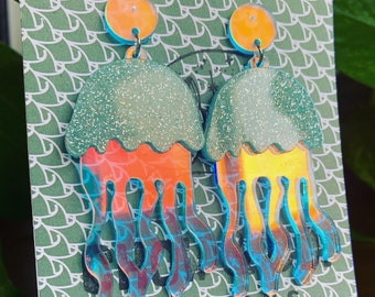 May include: A pair of jellyfish earrings with orange, teal, and iridescent accents. The jellyfish have a glittery teal top and orange and yellow bodies with teal tentacles. The earrings are hanging on a green patterned background. The word "CREATIONS" is printed at the bottom.