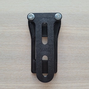 Op de afbeelding: Een zwarte, 3D-geprinte beugel met twee metalen bouten en moeren. De beugel heeft een rechthoekige vorm met een reeks sleuven en een gebogen bodem. De hardware is zilverkleurig. De beugel is ontworpen om een item vast te houden.