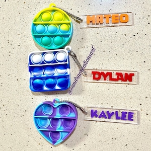 May include: Three silicone sensory keychains in circle, square, and heart shapes with tie-dye colors. Each keychain features a clear acrylic rectangular tag attached by a metal chain, displaying the names MATEO in orange, DYLAN in red, and KAYLEE in purple.