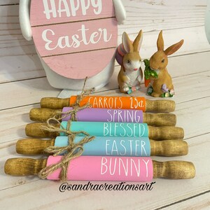 Puede incluir: Seis rodillos de amasar de madera pintados en colores pastel y con letras blancas. Los rodillos de amasar están dispuestos verticalmente con los mangos en la parte superior. Las letras en los rodillos de amasar dicen: Carrots, Spring, Blessed, Easter, Bunny y @sandrarcreationsart.