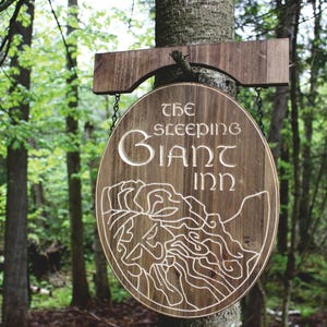 Op de afbeelding: Een rustiek houten bord voor "The Sleeping Giant Inn" hangt aan een boom in een bos. Het bord is rond met uitgesneden letters en een bergillustratie. Het bord hangt aan kettingen aan een houten beugel.