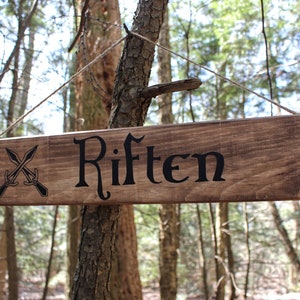 May include: A wooden arrow sign with the words "Riften" in black script. The sign is hanging from a tree branch in a forest. There are two crossed swords on the left side of the sign.