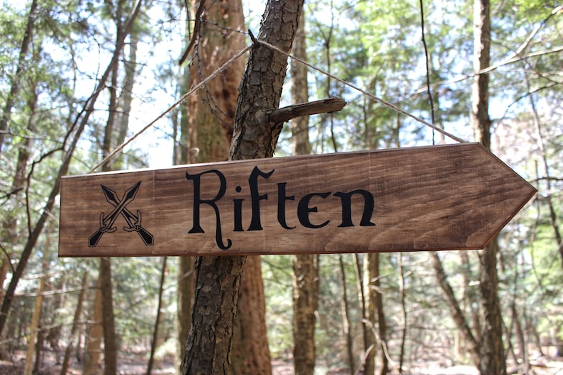 May include: A wooden arrow sign with the words "Riften" in black script. The sign is hanging from a tree branch in a forest. There are two crossed swords on the left side of the sign.