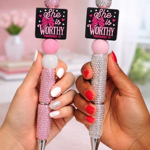 May include: Two decorative pens with rhinestone-covered barrels. One is pink, the other silver. Each pen has a black square with the text "She is Worthy" and a pink bow. The pens have a pink and white bead design.