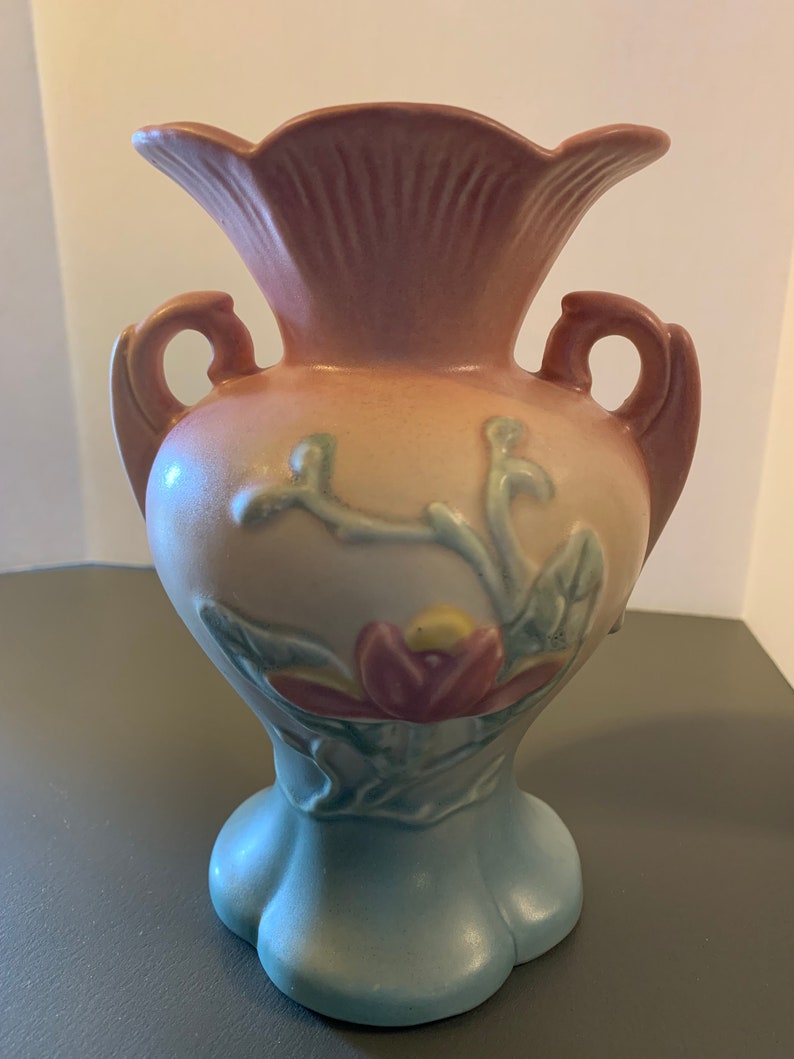 May include: A vintage ceramic vase with a pink, blue, and green glaze. The vase features a floral design with a large pink flower and green leaves. The vase has two handles and a flared opening.